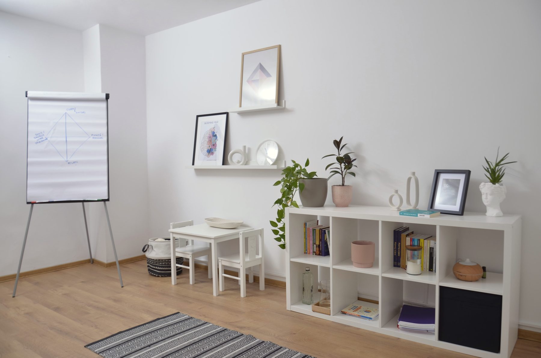 Sala de lucru cu flipchart, o măsuță pentru copii și un raft alb plin de cărți și plante