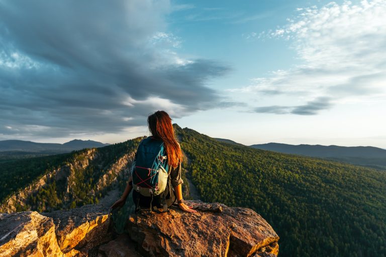 A young female tourist with a backpack admires the sunset from the top of the mountain